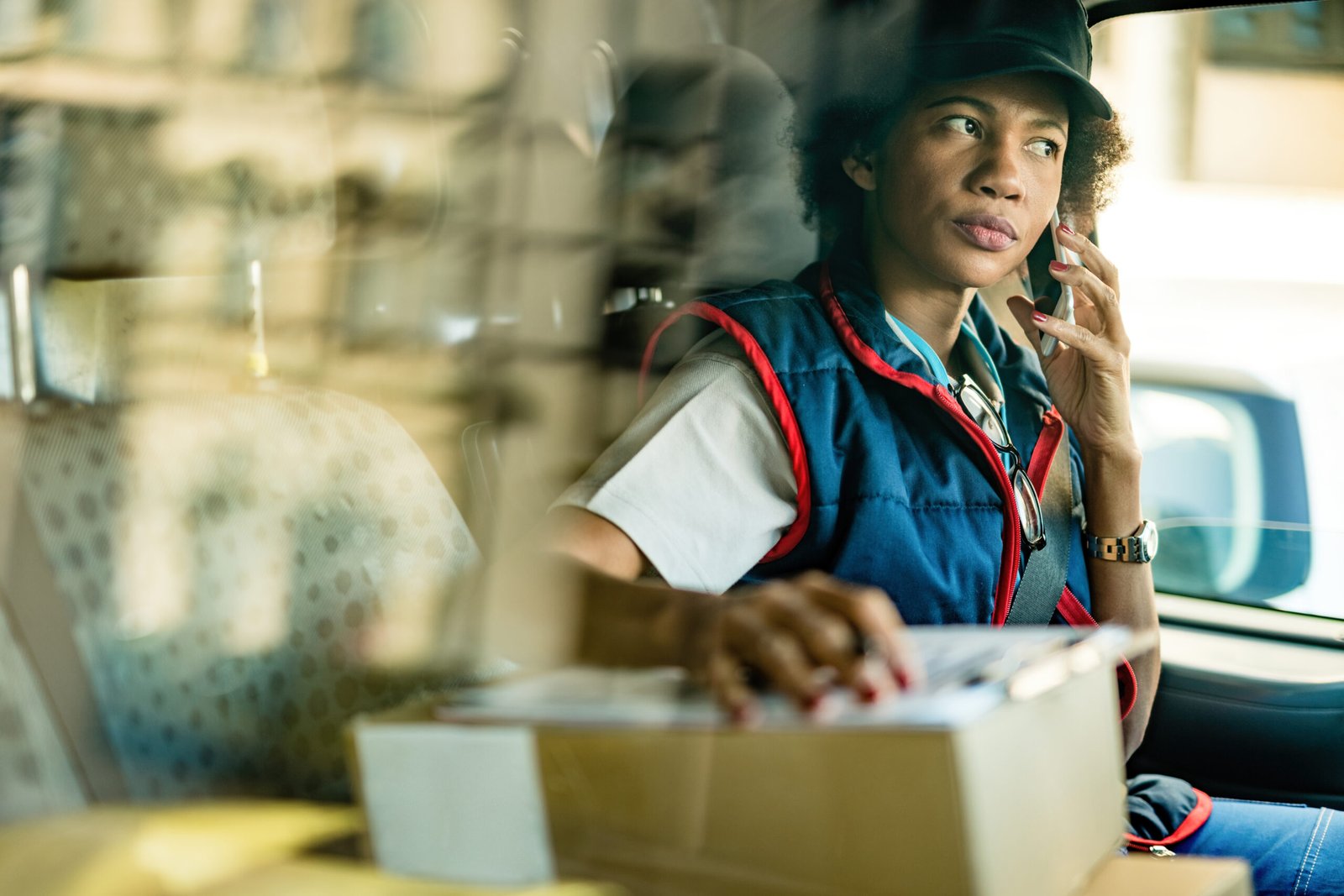 African American delivery woman talking with customer over mobile phone while sitting in a mini van. The view is through window.